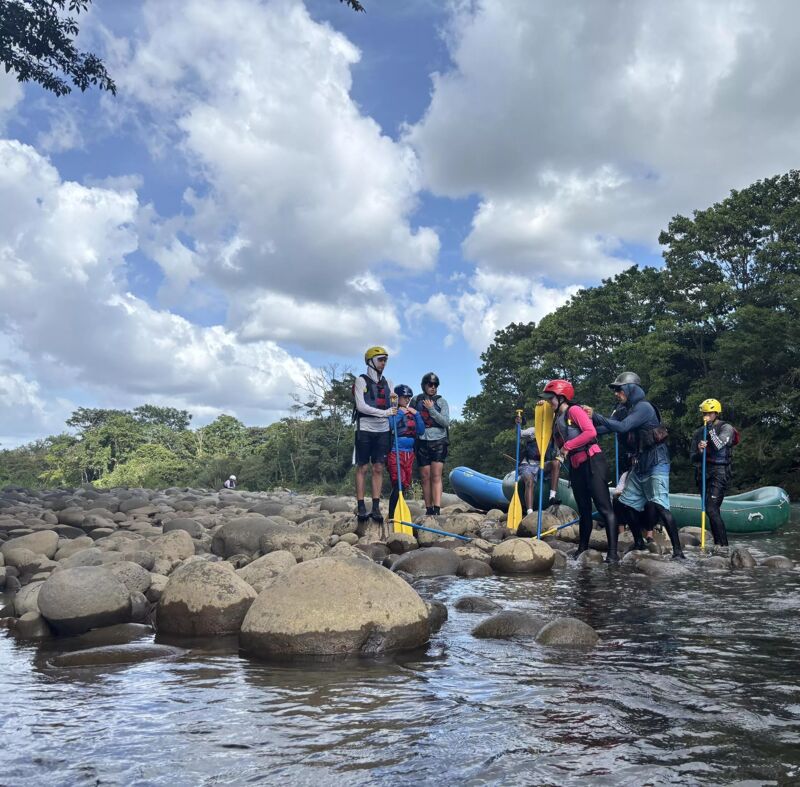 A group of people wearing life vests and helmets are preparing for a rafting adventure on a rocky river. They are standing near an inflatable raft, with paddles in hand, ready to navigate the river's currents. The sky is filled with fluffy clouds, and lush greenery lines the riverbank, creating a scenic backdrop for their outdoor activity.